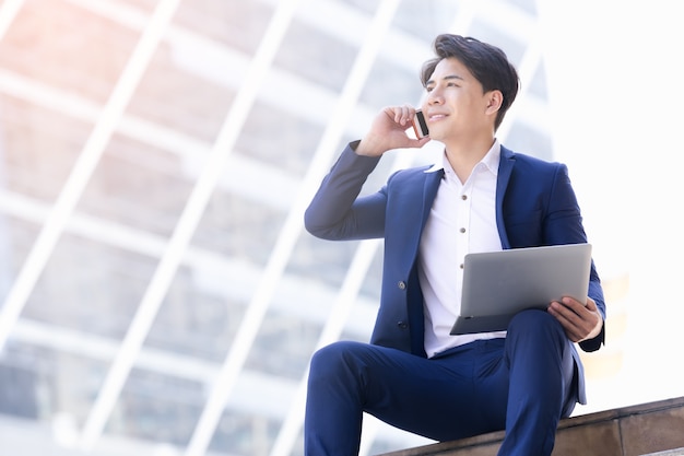 La sonrisa asiática del hombre de negocios trabaja la relajación feliz con la computadora portátil en ciudad