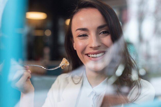 Sonrisa blanca como la nieve. Mujer sincera atractiva alegre que sonríe mientras que lleva tenedor y que presenta en el fondo borroso