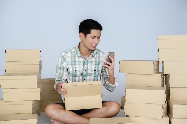 La sonrisa y la cara feliz del hombre asiático miran el teléfono inteligente con su tienda en línea entre muchas cajas con paquetes en una pared blanca. Concepto de inicio independiente y oficina en casa de negocios en línea.