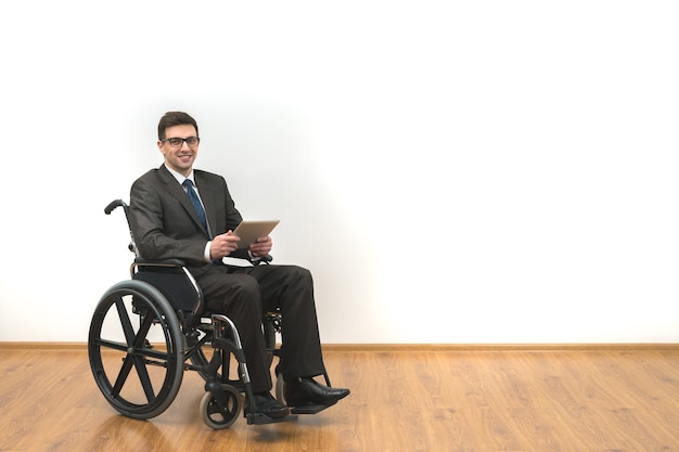 La sonrisa discapacitada en la silla de ruedas sosteniendo una tableta sobre un fondo de pared blanca