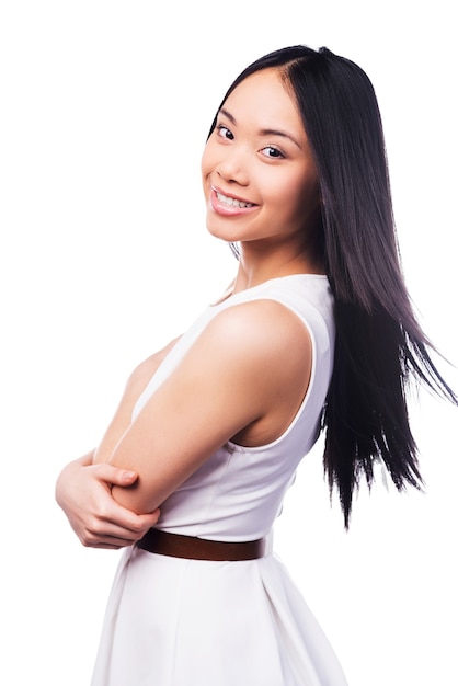 ¡Esta sonrisa es solo para ti! Hermosa joven asiática en vestido bonito manteniendo los brazos cruzados y sonriendo mientras está de pie contra el fondo blanco.