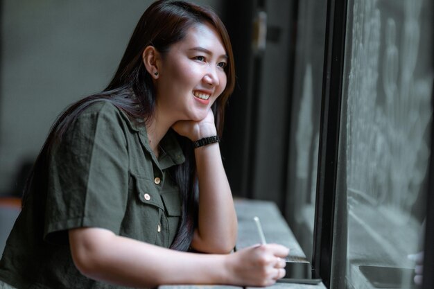 Sonrisa feliz, gente asiática independiente, mujer de negocios, trabajo informal, tomando nota en una tableta digital con bolígrafo electrónico, con un vestido verde pardusco, hipster elegante, Verano en una cafetería, Fondo de café