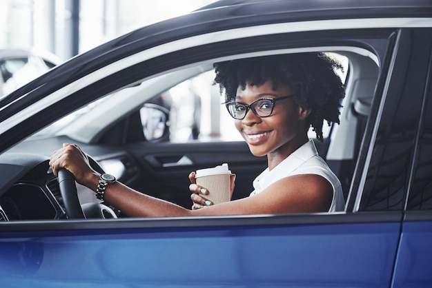 Sonrisa feliz. Joven mujer afroamericana se sienta dentro del coche nuevo y moderno.