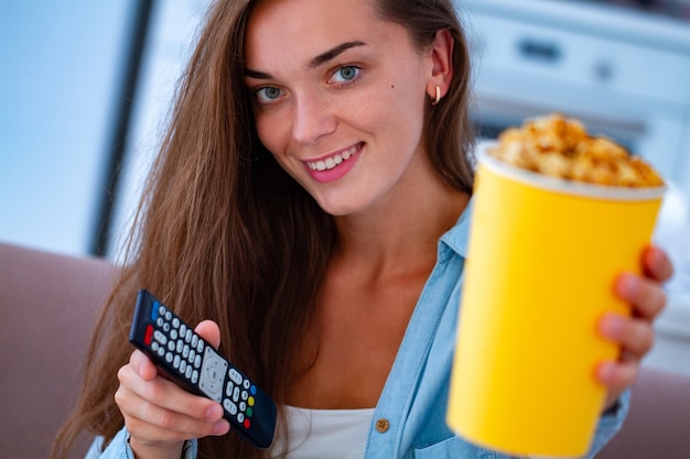 Sonrisa feliz mujer sosteniendo crujiente caja de palomitas de caramelo y control remoto de TV durante la televisión en casa