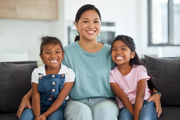 Sonrisa feliz y retrato de una madre y sus hijos sentados en un sofá en casa en Indonesia Felicidad amor y mamá abrazando a sus niñas mientras se relaja en un sofá juntos en su sala de estar