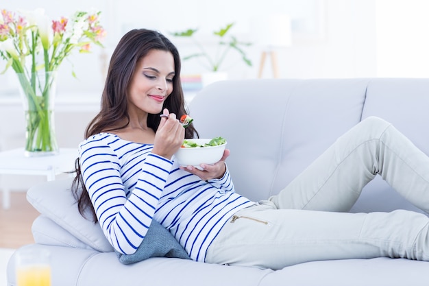 Sonrisa hermosa morena relajándose en el sofá y comiendo ensalada