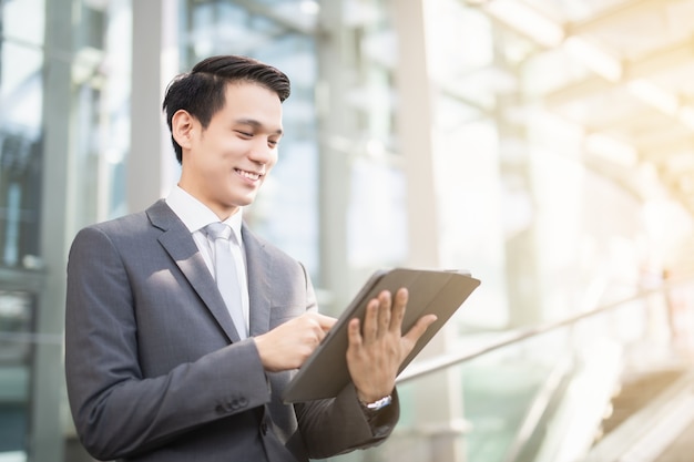 Sonrisa de hombre de negocios asiático y de pie en la oficina principal durante el uso de un stock de verificación de computadora portátil