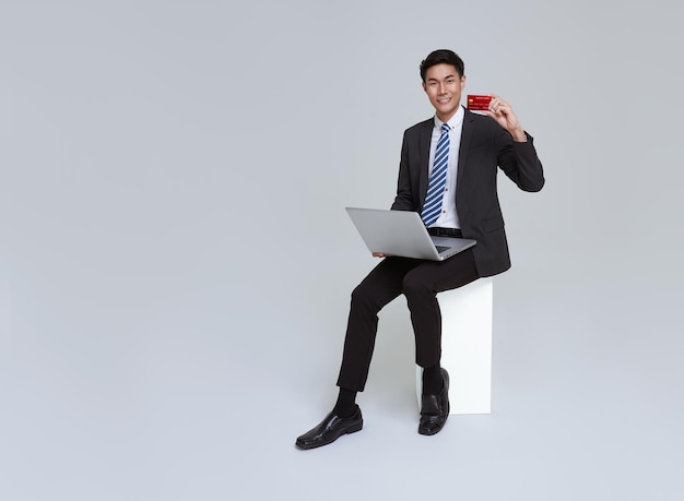 Sonrisa de hombre de negocios asiático en traje formal sentado en una silla usando una computadora portátil y mostrando la tarjeta de crédito