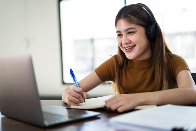 Sonrisa joven estudiante asiática usa auriculares inalámbricos, escriba en el cuaderno para estudiar el idioma en línea, vea y escuche al conferencista, seminario web a través de videollamadas, e-learning en el hogar, educación a distancia