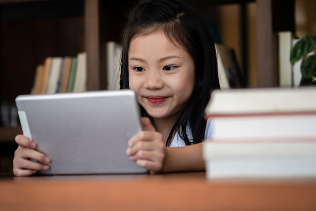 Sonrisa linda chica sentarse y jugar tableta inteligente en la biblioteca