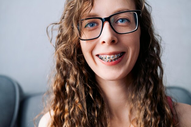 Foto sonrisa de una mujer joven de cabello largo con gafas y ojos azules en aparatos ortopédicos de primer plano