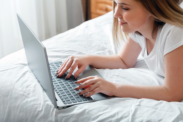 Sonrisa de mujer joven escribiendo en el teclado de la computadora portátil mientras está acostado en la cama en casa. La muchacha adolescente feliz se comunica en línea trabajando a distancia, estudiando compras o planificando su computadora portátil de uso diario en el dormitorio.