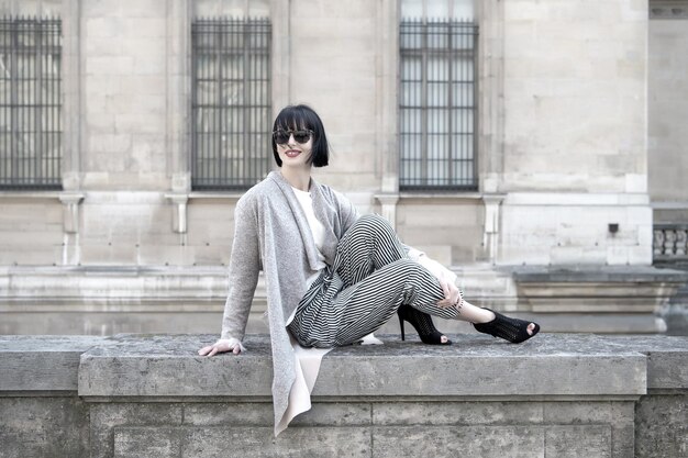 Sonrisa de mujer con labios rojos en gafas de sol, zapatos de tacón, ropa de moda en la valla de piedra en la fachada de la casa. Moda, belleza, concepto de estilo, mujer parisina