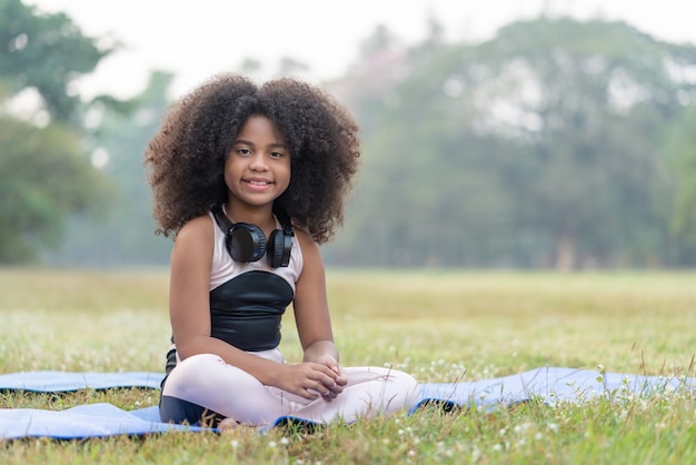 Sonrisa de niña afroamericana y sentada en la estera enrollable practicar yoga de meditación en el parque al aire libre