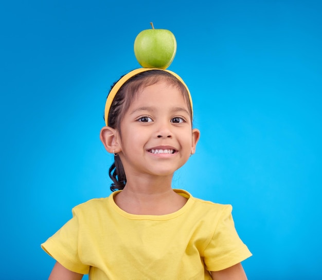 Sonrisa de retrato y niña con manzana en la cabeza sobre fondo azul para nutrición alimentación saludable y dieta Juventud de alimentos y cara de niña feliz en estudio con fruta para vitaminas orgánicas y bienestar