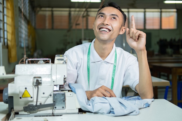 Sonrisa de sastre masculino asiático con la mano apuntando hacia arriba para presentar algo al coser con la máquina de coser