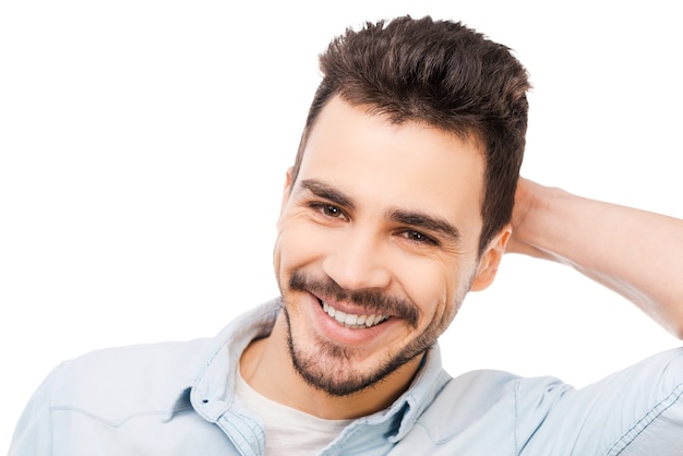 Sonrisa sincera. Apuesto joven tocando su cabeza y sonriendo mientras está de pie contra el fondo blanco.