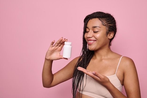 Sonrisas de mujer atractiva apuntando a un recipiente blanco con suplementos dietéticos o vitaminas para una piel y cabello saludables. Fondo rosa, espacio de copia. Concepto de tratamiento de salud, medicina y belleza.