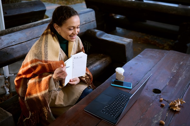 Sonrisas de una mujer bonita que se muestran con un bolígrafo en una página en blanco con una lista de tareas pendientes durante la videoconferencia en la computadora portátil, se mantiene caliente envolviéndose en una manta de lana a cuadros, sentada al aire libre en un banco de madera