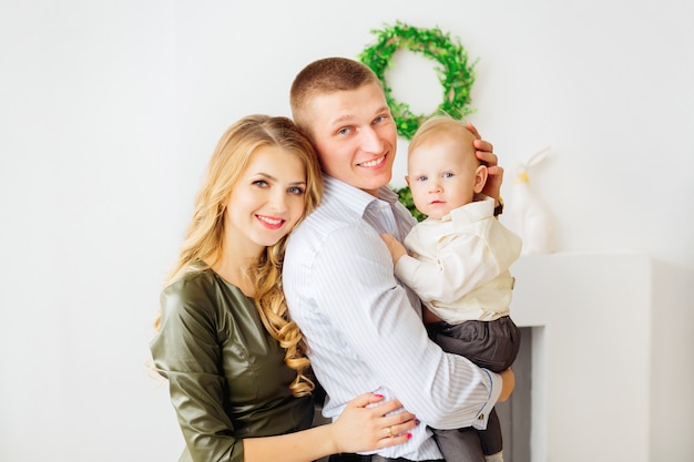 Sonrisas sinceras de padres que sostienen en las manos a su pequeño hijo contra el fondo de una chimenea y una corona verde