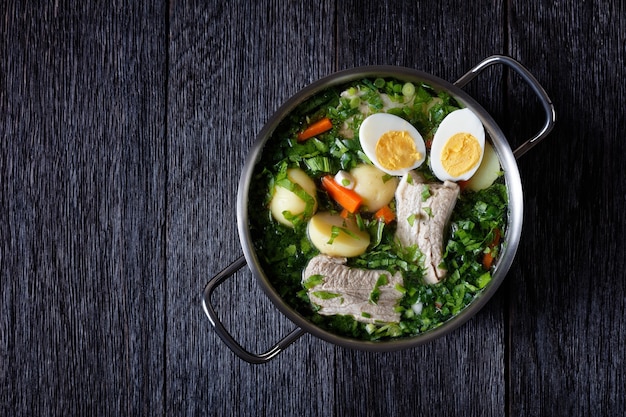 Sopa de acedera verde con perejil y cebolla verde con costillas de cerdo, con caldo de carne y mitades de huevo duro, servido en una olla de metal sobre una mesa de madera oscura, vista desde arriba, espacio de copia