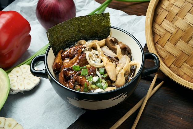 Sopa asiática clásica con fideos udon, nori, hongos shiitake y pollo frito al wok en un tazón negro sobre una mesa de madera con espacio de copia. Vista superior, comida plana