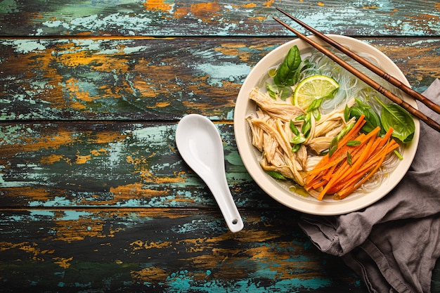 Sopa asiática com macarrão de arroz, frango e legumes em uma tigela de cerâmica servida com colher e pauzinhos em um fundo de madeira rústico de cima com espaço para texto, cozinha chinesa ou tailandesa