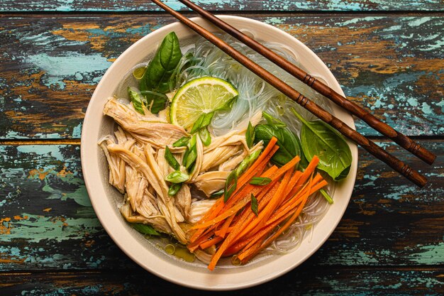 Sopa asiática com macarrão de arroz, frango e legumes em uma tigela de cerâmica servida com colher e pauzinhos em um fundo rústico de madeira de cima, cozinha chinesa ou tailandesa