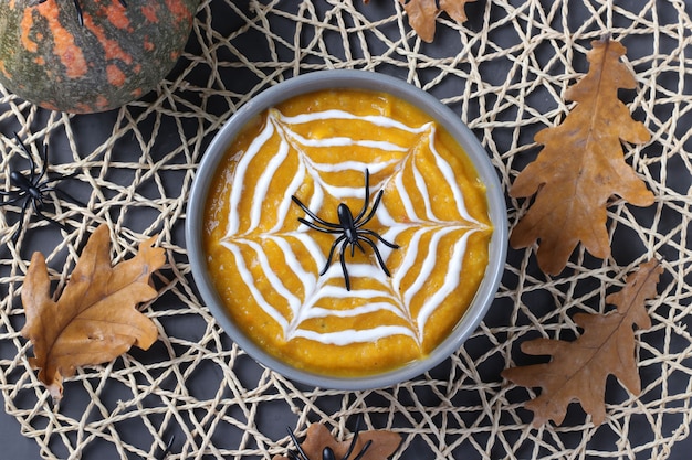 Sopa de calabaza de Halloween con telaraña cremosa en tazón gris y arañas sobre la mesa. Vista desde arriba.