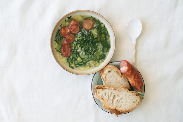 Sopa de Caldo Verde con verduras y chorizo picado por encima en tazón de cerámica con cuchara de cerámica. Trozos de pan y chorizo en plato de cerámica. Vista superior