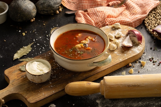 Sopa caseira com bolinhos, creme de leite, cebola e especiarias. Uma tigela de sopa, uma colher, uma toalha, um rolo de massa e ingredientes em uma mesa preta.