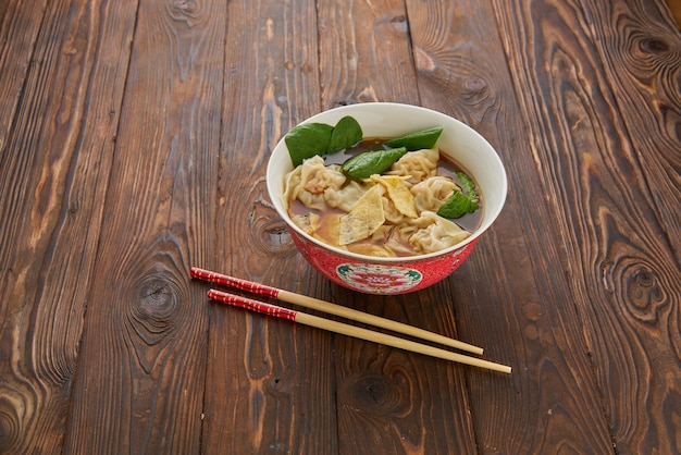 Sopa caseira de bolinho de massa wonton asiático na tigela tradicional chinesa vermelha com pauzinhos de madeira sobre a mesa de textura de madeira. Vista de cima plana, cópia espaço conceito de comida da Ásia