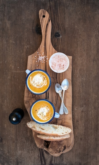 Sopa caseira de creme de abóbora em canecas de esmalte com ervas e fatias de pão fresco na azeitona, servindo a placa sobre a mesa de fundo de madeira rústica
