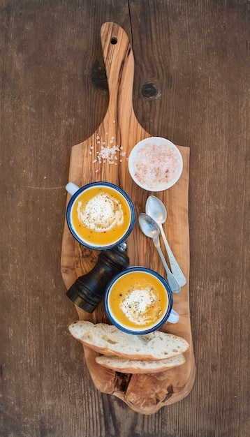 Sopa casera de crema de calabaza en tazas de esmalte con hierbas y rebanadas de pan fresco en una tabla de servir de aceitunas