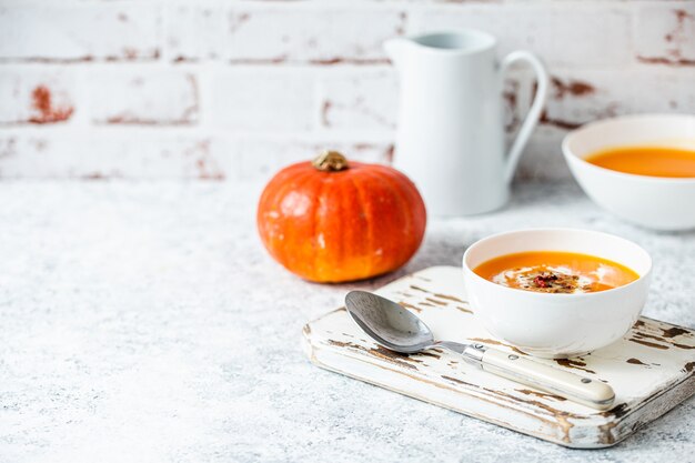 Sopa de crema de calabaza casera servida en cuenco de cerámica blanca sobre mesa blanca con cuchara decorada con calabaza entera, ángulo de visión, enfoque selectivo. Comida reconfortante acogedora de otoño, espacio de fondo para texto