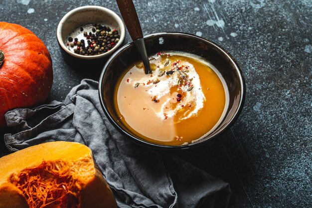 Sopa de crema de calabaza casera servida en cuenco de cerámica marrón sobre fondo de piedra oscura con cuchara decorada con calabaza fresca cortada, vista superior. Comida reconfortante tradicional de otoño