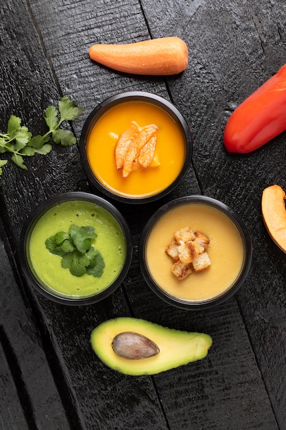 Sopa de crema de espinacas, sopa de calabaza y aguacate en recipientes de comida, plano. Fondo de madera, vertical