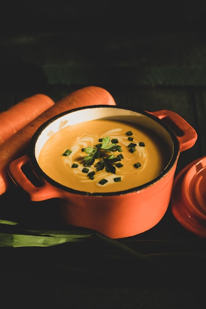 Sopa de crema de zanahoria en cazuela de madera verde oscuro con tapa naranja con dos zanahorias frescas más cebollino