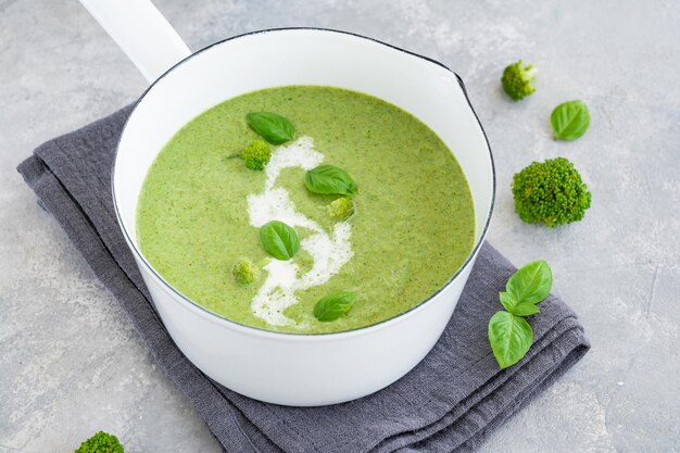 Sopa creme de brócolis com manjericão fresco e em uma panela branca sobre fundo cinza de concreto