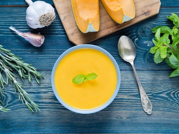 Sopa cremosa de calabaza sobre fondo de madera azul oscuro, romero, ajo, menta, rodajas de p