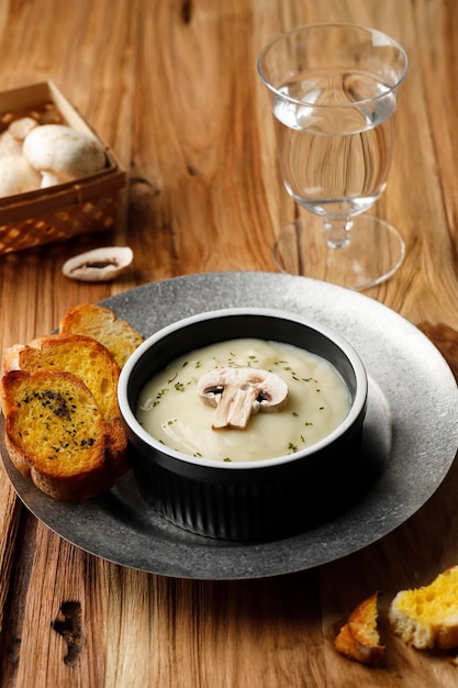 Sopa cremosa de champiñones con baguette de pan de ajo crujiente, servir en un tazón de cerámica negra o ramequín, en una mesa rústica de madera marrón.