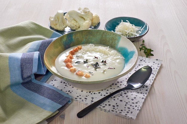 Sopa cremosa de coliflor en una taza azul con crutones, tomillo y queso parmesano sobre madera clara
