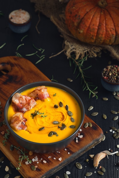 Sopa cremosa de abóbora com fatias de bacon salteadas com creme e sementes de abóbora em uma tigela preta sobre uma mesa de madeira preta. vista superior, foco seletivo. ingredientes para fazer sopa de abóbora sazonal na mesa