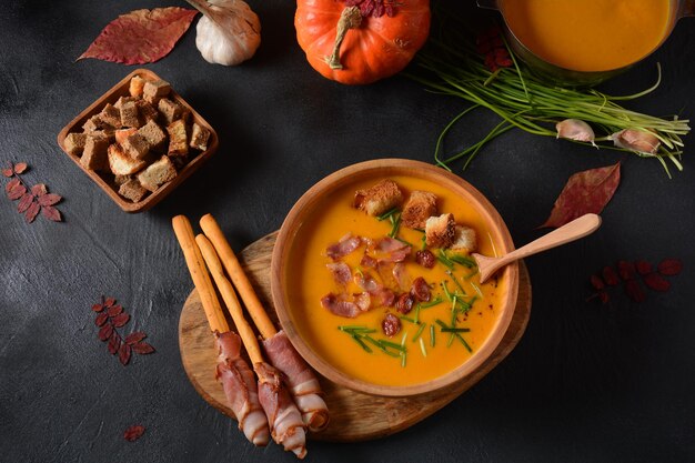 Sopa cremosa de abóbora e batata doce (batata) com bacon em fundo escuro. Uma tigela de sopa com croutons caseiros e ervas com decorações de outono.