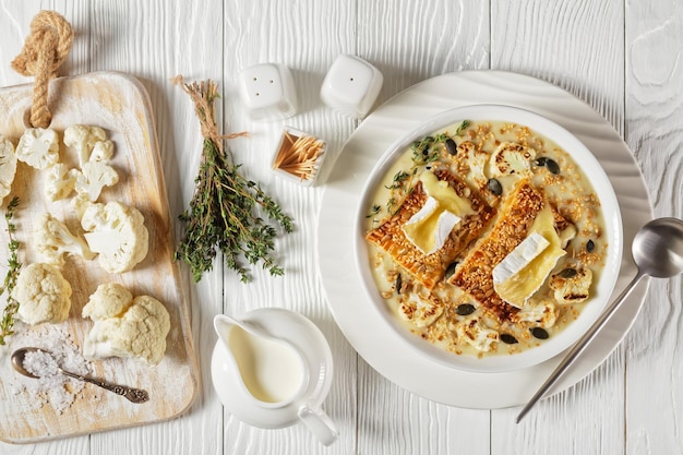 Sopa cremosa de couve-flor vegana coberta com torradas com queijo camembert derretido em uma tigela branca sobre uma mesa de madeira com ingredientes, vista horizontal acima, close-up, flat lay, cozinha francesa