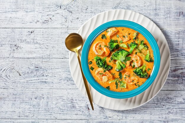 Sopa cremosa de tomate con camarones y brócoli en un recipiente azul sobre una mesa de madera blanca, cocina americana