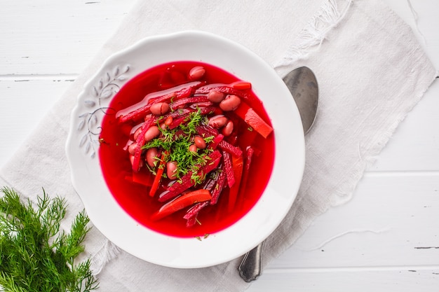 Sopa da beterraba do vegetariano com feijões e vegetais na placa branca no fundo de madeira branco.