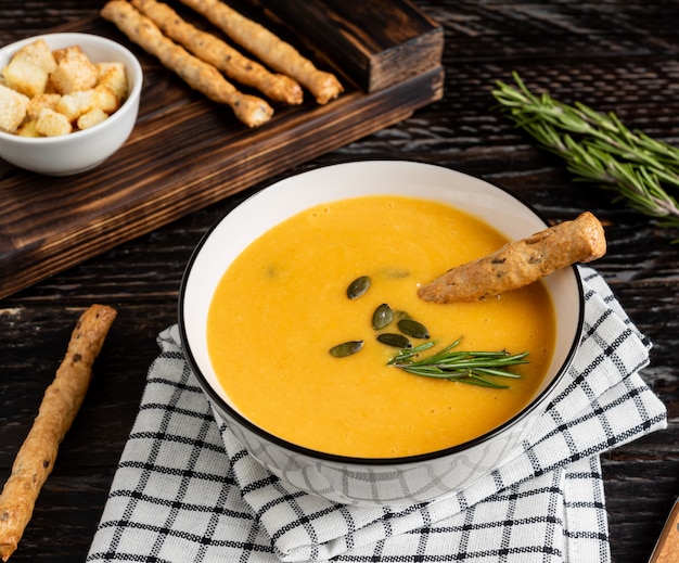 Sopa de abóbora e cenoura com creme, sementes de abóbora, croutons e pão grossini varas no fundo escuro de madeira.