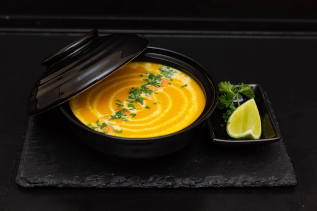 Sopa de abóbora em um prato preto sobre um fundo preto de madeira ao lado de uma fatia de limão