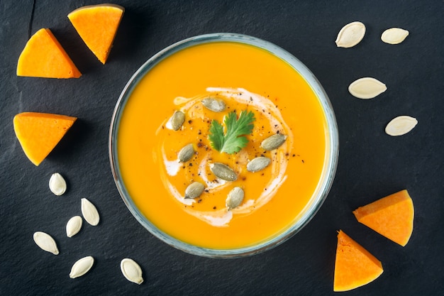 Sopa de abóbora em uma tigela. Fatias e sementes da polpa de Butternut na placa da ardósia. Comida vegetariana saudável. Dia das bruxas, jantar de ação de Graças. Copie o espaço.